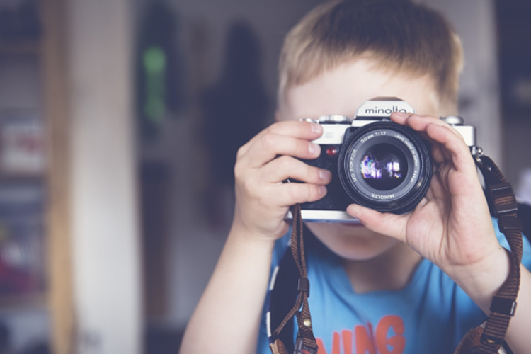 Postać dziecka w wieku ok 7 lat, robiącego zdjęcie obserwatorowi, tradycyjnym aparatem fotograficznym