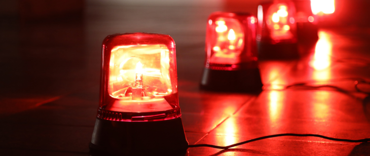 Na obrazku widać kilka czerwonych lamp ostrzegawczych (sygnałowych) ustawionych na podłodze i połączonych przewodami. Lampy świecą intensywnym czerwonym światłem, a jedna z nich jest na pierwszym planie, podczas gdy pozostałe są lekko rozmyte w tle. Możliwe, że służą jako oświetlenie alarmowe lub sygnalizacyjne.