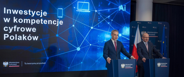 Na obrazku widać konferencję prasową Ministerstwa Cyfryzacji. Dwóch mężczyzn stoi przy mównicach z mikrofonami, a w tle znajduje się duży ekran z napisem „Inwestycje w kompetencje cyfrowe Polaków” oraz grafiką sieci i urządzeń cyfrowych. Obok widoczne są flagi Polski i Unii Europejskiej.