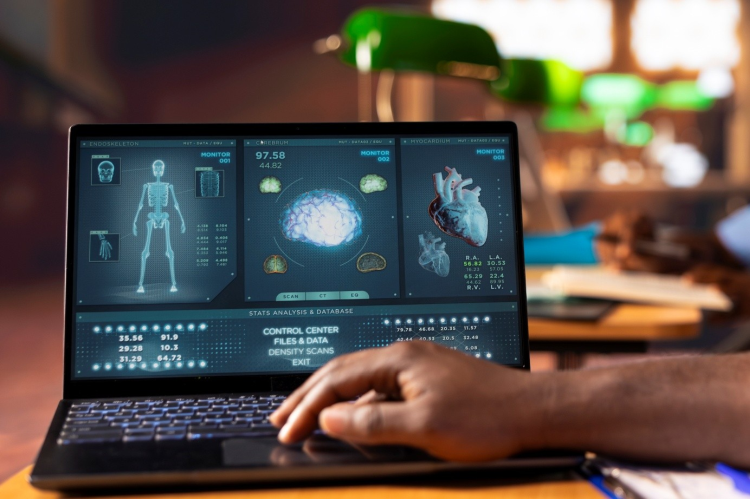 Dłoń obsługuje laptop z ekranem pokazującym medyczne wizualizacje ciała, mózgu i serca oraz dane diagnostyczne; zdjęcie sugeruje analizę wyników badań lub nowoczesny system medyczny.