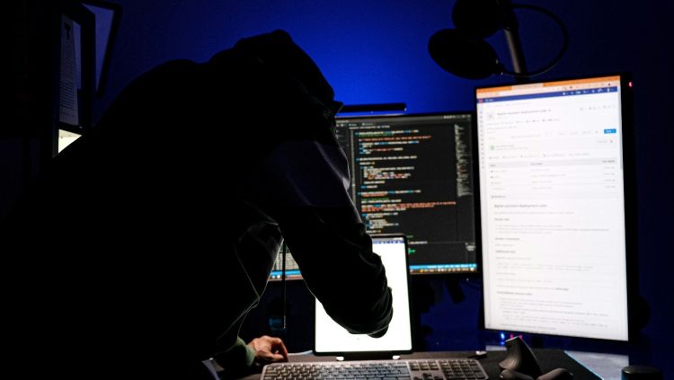 Obraz przedstawia osobę pracującą przy kilku monitorach komputerowych, na których widoczny jest kod oraz interfejsy systemowe. Ciemna kolorystyka i anonimowa sylwetka nawiązują do tematyki cyberbezpieczeństwa, cyberataków i działalności hakerskiej.