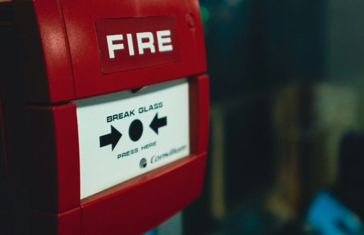 Obraz przedstawia ręczny przycisk alarmu pożarowego w czerwonej obudowie, oznaczony napisem „FIRE”. Urządzenie służy do ręcznego uruchomienia alarmu w przypadku zauważenia pożaru i stanowi element systemu bezpieczeństwa przeciwpożarowego.